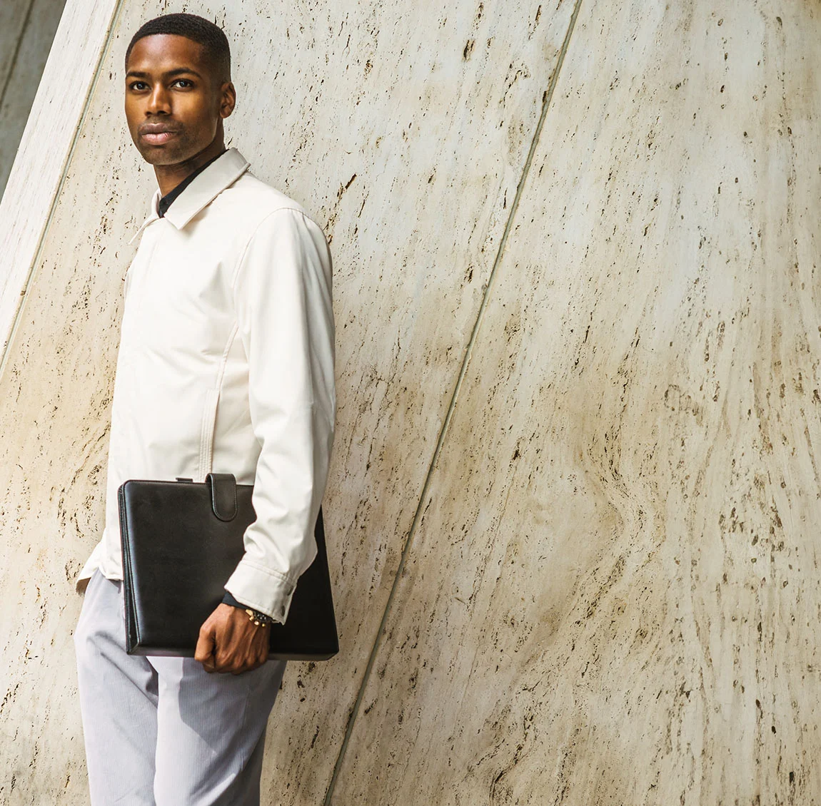 Stylish man with short hair wearing a light jacket and holding a black leather portfolio, standing against a marble wall - Functional Rhinoplasty at HIM Plastic Surgery